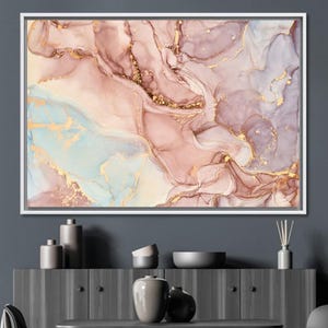 May include: Abstract art print in a white frame. The artwork features a fluid design with shades of pink, gold, and blue. The colors blend together to create a marbled effect, with gold accents throughout the design. The artwork is displayed on a dark gray wall.