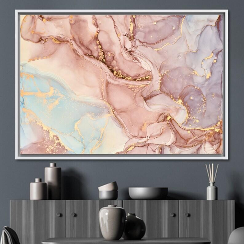 May include: Abstract art print in a white frame. The artwork features a fluid design with shades of pink, gold, and blue. The colors blend together to create a marbled effect, with gold accents throughout the design. The artwork is displayed on a dark gray wall.