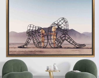 Op de afbeelding: Een ingelijste kunstdruk met een surrealistische sculptuur van twee figuren in een woestijnlandschap. De figuren zijn draadframe menselijke vormen, elk met een kleinere, goudkleurige figuur. Het kunstwerk is geplaatst tegen een achtergrond van bergen en een bleke lucht.