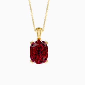 Può includere: Una collana con pendente color oro con una grande pietra preziosa rosso scuro taglio cuscino. La pietra preziosa è tenuta in posizione da quattro griffe dorate e pende da una delicata catena d'oro. Il pendente è centrato su sfondo bianco.