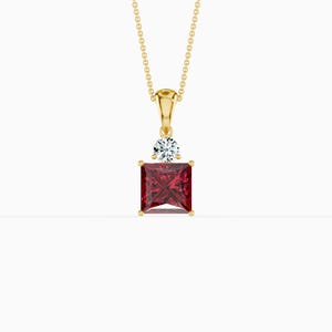 May include: A gold-colored pendant necklace featuring a square, deep red gemstone. A small, round, clear gemstone sits above the square stone. The necklace chain is also gold-colored.