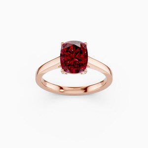 May include: A rose gold ring featuring a large, cushion-cut, deep red gemstone. The gemstone is the focal point, set in a four-prong setting, with a smooth, polished band. The ring is displayed against a white background.