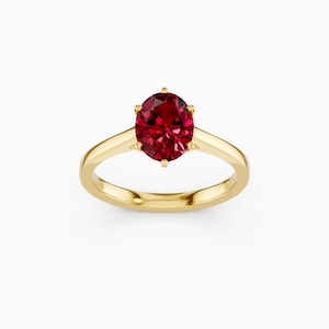 Pode incluir: Um anel de ouro com uma grande pedra preciosa vermelha escura em forma oval. A pedra é presa por seis garras. A banda do anel é de ouro polido simples.