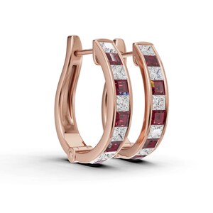 May include: A pair of rose gold hoop earrings. Each earring has a row of square-cut, alternating red and white gemstones. The earrings have a hinged closure and are shown against a white background.