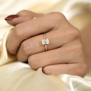 以下が含まれることがあります： 長方形のエメラルドカットダイヤモンドをあしらった、ローズゴールドのリング。リングは繊細なバンドにセットされています。リングは、ニュートラルな背景を持つ手の上に表示されています。