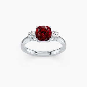 Puede incluir: Un anillo plateado con una gran gema roja oscura de talla cojín en el centro, flanqueada por dos gemas redondas y claras más pequeñas. La banda del anillo es lisa y pulida, creando un diseño clásico y elegante.