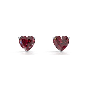 May include: A pair of heart-shaped, red gemstone stud earrings. The gemstones are a deep red color and are set in a silver metal. The earrings are displayed against a white background.