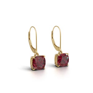 May include: A pair of gold-colored leverback earrings, each featuring a square, deep red gemstone. The gemstones are set in a gold bezel, and the earrings are displayed against a white background.
