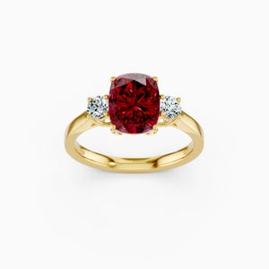 May include: A gold ring featuring a large, cushion-cut, deep red gemstone as the centerpiece, flanked by two smaller, round, clear gemstones. The ring's band is a polished gold color.