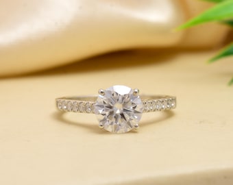 May include: A silver ring with a large, round, clear gemstone as the centerpiece. The band is encrusted with smaller, clear gemstones. The ring is set against a soft, neutral background, with a hint of green foliage in the corner.