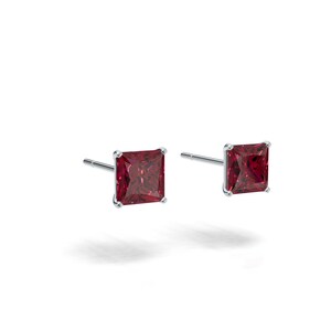 May include: A pair of stud earrings featuring square-cut, deep red gemstones set in a silver-colored metal. The earrings are displayed against a white background, showcasing their color and design. The gemstones have a faceted appearance.
