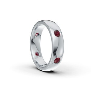 May include: A polished silver-toned band ring featuring a smooth, rounded surface with three small, circular, deep red gemstones embedded at regular intervals along the outer circumference of the metal band.