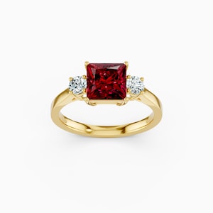 May include: A gold ring featuring a large, square-cut, deep red gemstone as the centerpiece, flanked by two smaller, round, clear gemstones. The ring's band is a polished gold color, creating a classic and elegant design.