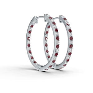 May include: A pair of silver hoop earrings. Each earring is encrusted with alternating red and white gemstones. The earrings are set against a white background, highlighting their sparkle and design. The gemstones are evenly spaced around the hoops.