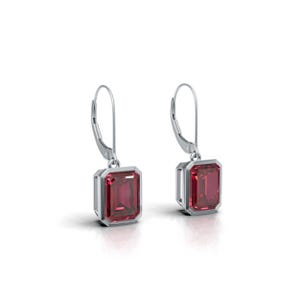 Pode incluir: Um par de brincos pendurados prateados. Cada brinco apresenta uma pedra preciosa retangular, vermelho escuro, emoldurada em prata. Fechamento por alavanca. Fundo branco.