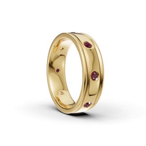 May include: A polished yellow gold band ring featuring a smooth, rounded surface with a central channel. The ring is set with multiple small, round, deep red rubies spaced evenly around the circumference of the band in a flush setting.