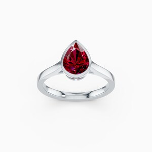 May include: A silver ring featuring a pear-shaped, deep red gemstone. The gemstone is set in a bezel setting, and the band is smooth and polished. The ring is displayed against a white background.