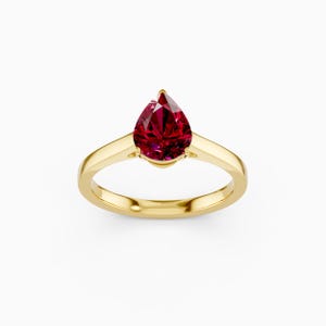 Può includere: Un anello d'oro con una gemma rosso scuro a forma di pera. L'anello ha una fascia in oro lucido e la gemma è tenuta da sottili griffe. Le sfaccettature della gemma riflettono la luce, creando un bagliore.