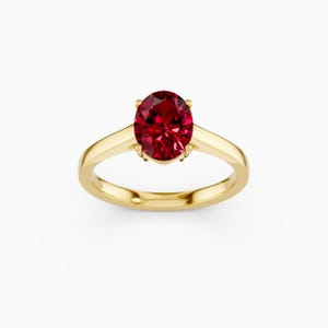May include: A gold ring featuring a large, oval-cut, deep red gemstone. The gemstone is the focal point, set in a classic four-prong setting. The band is a smooth, polished gold.