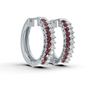 May include: A pair of silver hoop earrings. Each earring is adorned with alternating rows of small, round, red gemstones and clear, white gemstones. The earrings are set against a white background.