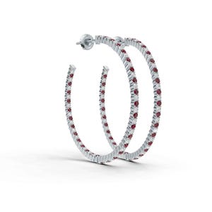 May include: A pair of silver hoop earrings, each adorned with alternating red and clear gemstones. The earrings are set against a white background, highlighting their sparkle and design. The earrings are a classic jewelry item.