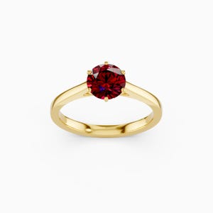 May include: A gold ring with a large, round, deep red gemstone. The gemstone is set with six prongs, and the band is a simple, polished gold. The ring is centered on a white background.