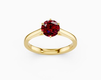 May include: A gold ring featuring a large, round, deep red gemstone. The gemstone is held in place by six prongs, and the band is polished gold. The ring is set against a white backdrop.