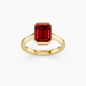 Puede incluir: Un anillo de oro con una piedra preciosa rectangular de color rojo intenso. La piedra preciosa está engastada en un bisel y el anillo tiene una banda lisa y pulida. El anillo se muestra sobre un fondo blanco.