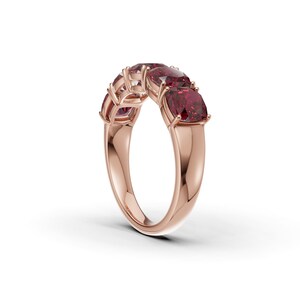Peut inclure: Une bague en or rose ornée de cinq pierres précieuses rouge foncé taille coussin disposées en rangée sur le dessus. L'anneau est lisse et poli, chaque pierre étant fixée par quatre griffes métalliques. Le design présente un profil incurvé.
