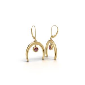 May include: A pair of gold drop earrings featuring a polished crescent moon shape with a small, round, bezel-set pink gemstone suspended from the centre of each arch. The earrings are attached to lever-back closures for secure wear.