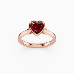 Könnte beinhalten: Ein roségoldener Ring mit einem herzförmigen, tiefroten Edelstein. Der Edelstein ist in einer Lünettenfassung gefasst, und das Band ist glatt und poliert. Der Ring wird vor einem weißen Hintergrund gezeigt.