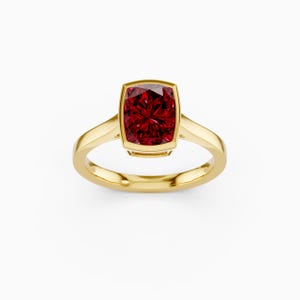 May include: A gold ring featuring a large, square-cut, deep red gemstone. The gemstone is set in a bezel setting, and the band is a simple, polished gold. The ring is displayed against a white background.