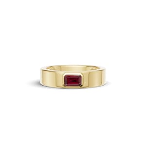 Può includere: Un anello a fascia in oro giallo lucido con una singola pietra preziosa di rubino rosso intenso dal taglio baguette rettangolare, incastonata orizzontalmente in una montatura a lunetta liscia al centro della fascia, dalla struttura solida.