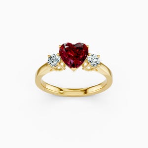 Può includere: Un anello d'oro con una pietra preziosa rosso scuro a forma di cuore al centro, affiancata da due pietre preziose rotonde e chiare più piccole. L'anello ha una fascia in oro lucido e un'incastonatura delicata.