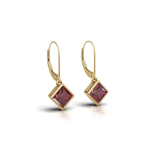 May include: A pair of gold-colored leverback earrings. Each earring features a square, deep red gemstone set within a gold bezel. The earrings are displayed against a white background, highlighting their design and color.