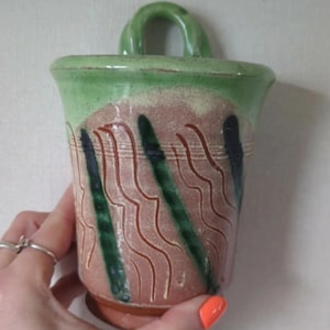 May include: Handmade ceramic wall pocket with a green handle and rim. The body has a pinkish-brown base with green and dark blue vertical stripes and wavy brown lines. A unique piece for home decor.