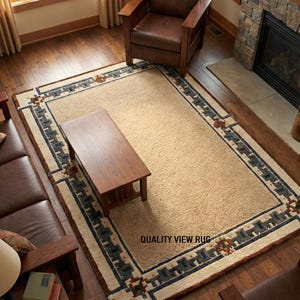May include: A rectangular beige rug with a geometric border in a living room setting. The border features a pattern of dark blue, brown, and cream-colored shapes. The rug is placed on a hardwood floor, with a leather chair and coffee table nearby. The text "QUALITY VIEW RUG" is visible.