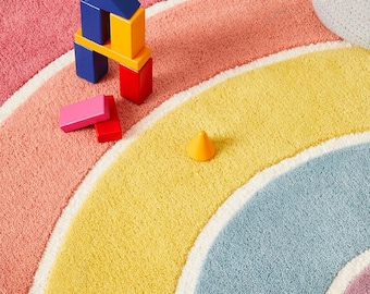 May include: A colorful, round rug with a rainbow design. The rug features bands of pink, orange, yellow, blue, and purple, separated by white borders. Wooden toy blocks in red, yellow, blue, and pink are scattered on the rug.