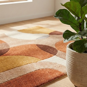 May include: A contemporary rug featuring a geometric design in orange, brown, beige, and yellow. The rug has a soft, textured surface. A potted plant with large green leaves is positioned beside it. The rug is rectangular.