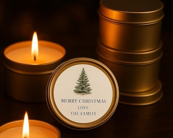 Puede incluir: Latas de velas doradas con llamas encendidas y una tapa decorativa. La tapa presenta una ilustración de árbol de Navidad y el texto "MERRY CHRISTMAS LOVE THE FAMILY". Una pila de latas doradas está en el fondo.