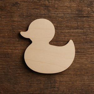 May include: A wooden duck-shaped cutout, light tan in colour with a smooth texture. The duck has a rounded head, a small beak, and a curved body. The cutout is placed on a dark brown wooden surface.