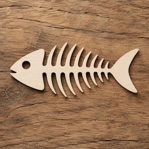 Puede incluir: Recorte de esqueleto de pez de madera, con cabeza, ojo y aleta caudal. El diseño de espina de pescado es de color marrón claro y descansa sobre una superficie de madera texturizada.