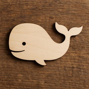 Puede incluir: Recorte de ballena de madera con una superficie lisa y de color claro. La ballena presenta un diseño sencillo con un ojo negro, una boca curva y una cola. El recorte es probablemente para manualidades o decoración.