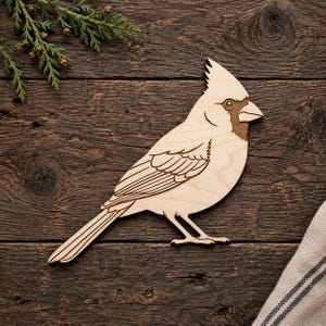 Op de afbeelding: Een platte, lasergesneden houten silhouet van een kardinaalvogel met gegraveerde vleugel- en veertjesdetails. De lichtgekleurde houten uitsparing toont een puntige kuif op de kop, een scherpe snavel en een lange staart met natuurlijke houtnerven.