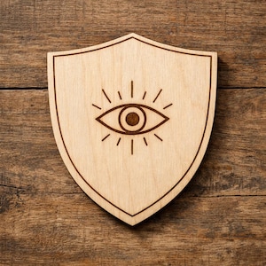 May include: A wooden shield with a light brown finish, featuring a detailed eye symbol in the center. The eye is surrounded by radiating lines, and the shield has a decorative border. The design is carved into the wood.