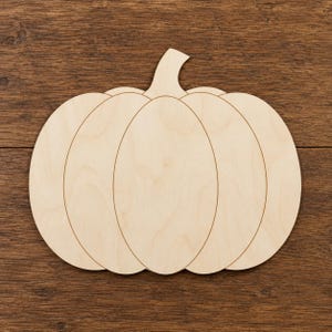 May include: A flat, light-coloured wooden cutout in the shape of a pumpkin. The object features a smooth, natural wood grain surface with clearly defined, laser-cut lines that delineate the pumpkin's segments and a small stem at the top centre.