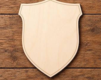 May include: A light-coloured wooden plaque shaped like a heraldic shield. The plaque features a smooth, flat surface with a natural wood grain pattern and a thin, dark engraved line following the contour of the shield's perimeter edge.