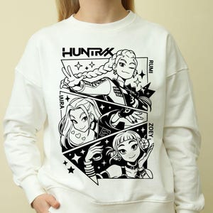 May include: White sweatshirt featuring a black and white graphic design with the word "HUNTRAX" at the top. The graphic includes three stylized female characters named Laira, Rumi, and Zoey, with star and geometric accents.