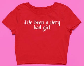 May include: A red crop top with the text "I've been a very bad girl" in white gothic font. The shirt has short sleeves and a round neckline. The background is a solid pink color.