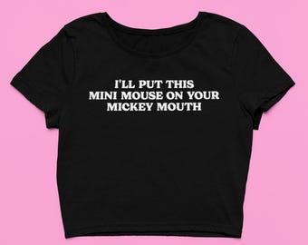 May include: Black crop top with the text "I'LL PUT THIS MINI MOUSE ON YOUR MICKEY MOUTH" in white. The shirt has short sleeves and a scoop neck. The background is a solid pink.